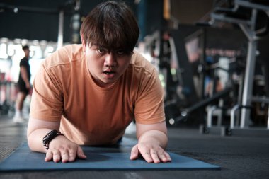 Kilolu adam spor salonundaki paspasın üzerinde tahta egzersizi yaparken kararlı. Fitness zorluğu, kilo kaybı ve sağlıklı yaşam tarzı kavramı.