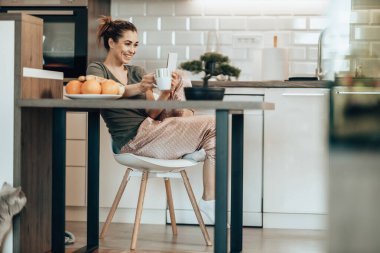 Genç, gülümseyen bir kadın sabah evde kahve içerken akıllı telefonunu kullanıyor..