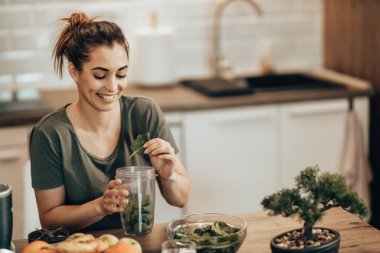 Young smiling woman putting a variety of  vegetables into a blender and making a healthy smoothie at home.