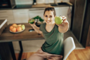 Shot of a smiling woman holding a green apple and glass of smoothie. Selective focus on an apple.