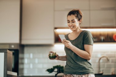 Genç gülümseyen kadın sabah evde yeşil smoothie içerken akıllı telefonunu kullanıyor.