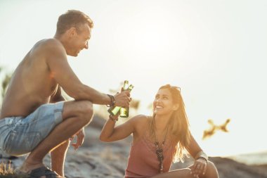 Young couple having fun and drinking beer on the beach by the sea. Enjoying in beautiful sunset and their love.