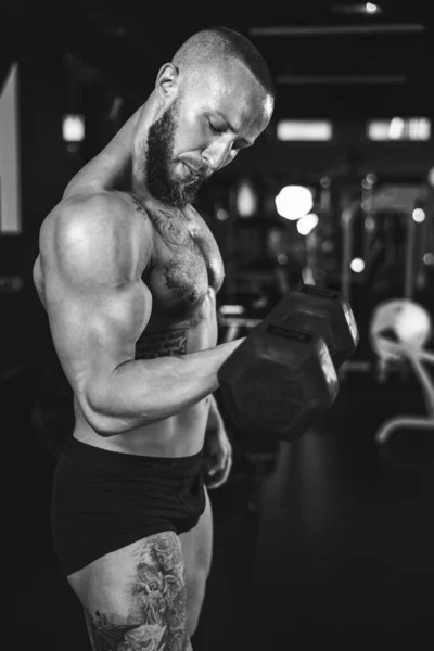 Black and white photo of a muscular bodybuilder doing hard training with dumbbell at the gym. He is pumping up his biceps muscle with heavy weight.