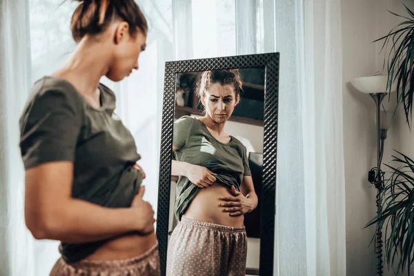 Frown young woman standing in front of a mirror and holding hands on her bloating stomach.