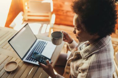 Siyah kadın sabah çayı içiyor ve tatilini rahat ahşap bir evde geçirirken akıllı telefon kullanıyor..