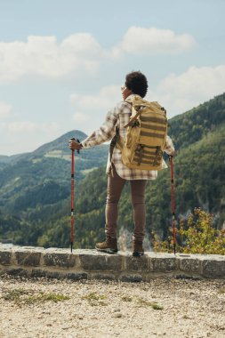 Sırt çantası ve yürüyüş direkleri olan siyah bir kadının arka görüntüsü doğada duruyor ve dağda yürüyüş yaparken temiz havanın tadını çıkarıyor..