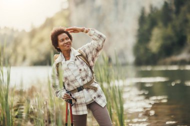 Sırt çantalı olgun siyah bir kadın nehir manzarasının keyfini çıkarıyor ve dağda yürüyüş yaparken dinleniyor..