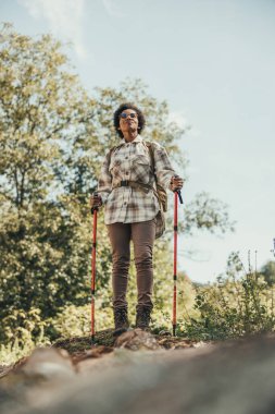 Olgun siyah kadın dağda yürüyüş sırıklarıyla gezerken manzaranın tadını çıkarıyor..