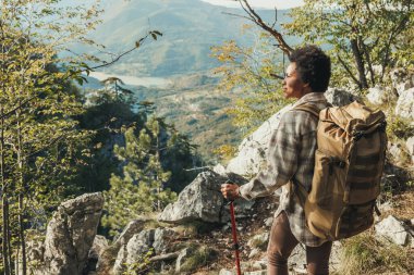 Olgun siyahi kadın bir tepenin üstünde duruyor ve dağlardaki yürüyüşü sırasında manzaranın tadını çıkarıyor..