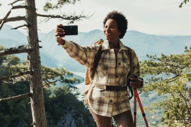 Yetişkin siyahi bir kadın bir tepede dikilirken ve dağlardaki yürüyüşü sırasında manzaranın tadını çıkarırken bir video görüşmesi yapıyor..