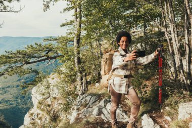Yetişkin siyahi bir kadın, bir tepenin zirvesinde dikilirken ve dağlardaki yürüyüşü sırasında manzaranın tadını çıkarırken selfie çekiyor..