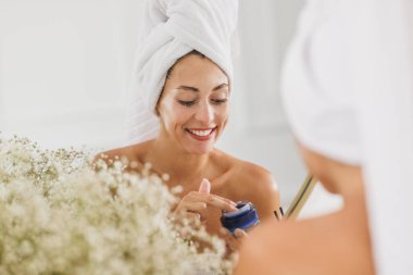 Güzel genç bir kadın aynanın karşısına geçip evdeki banyoda yüzüne nemlendirici sürüyor..