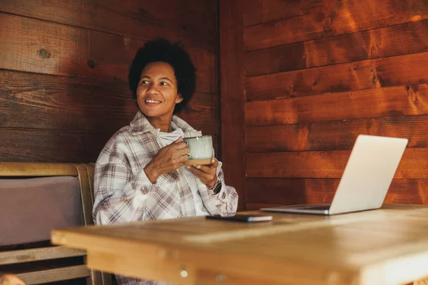 Siyah bir kadın sabah çayı içip dizüstü bilgisayar kullanırken, tatilini rahat, ahşap bir evde geçiriyor..