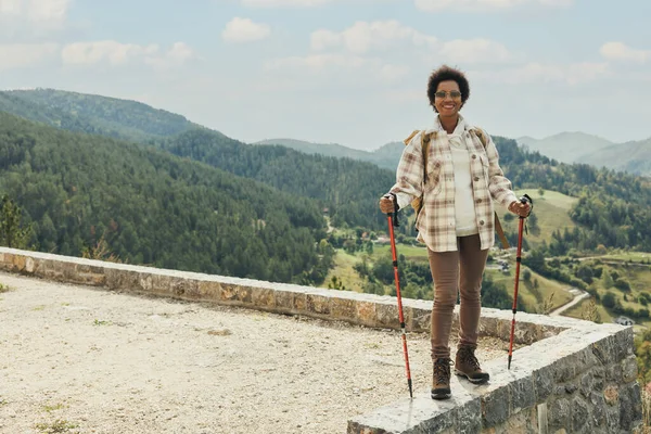 Mature black woman with backpack and trekking poles standing in nature and enjoying in fresh air while hiking on mountain.