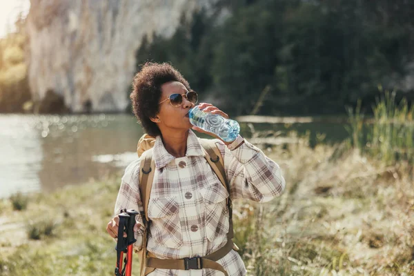 山の中でハイキング中に湖で水休憩を取るリュックを持つ熟女黒人女性 — ストック写真