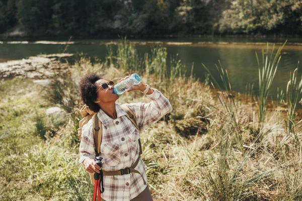 山の中でハイキング中に湖で水休憩を取るリュックを持つ熟女黒人女性 — ストック写真