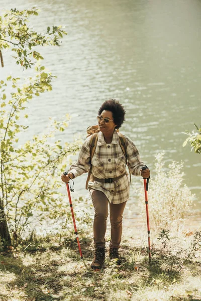Mature black woman with backpack and trekking poles taking a break by the lake while hiking.