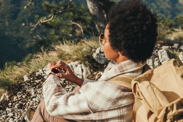 山の崖の上に座っている間 ビューを楽しんで成熟した黒人女性と彼女のハイキング冒険中にスマートウォッチを見て — ストック写真