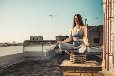 Rahat bir kadın terasta yoga yaparken meditasyon yapıyor. Arka planda şehir binaları.
