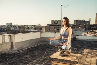 Rahat bir kadın terasta yoga yaparken meditasyon yapıyor. Arka planda şehir binaları.