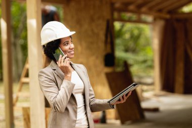 Beyaz kasklı, akıllı telefon ve dijital tabletle yeni ahşap bir evin şantiyesini kontrol ederken Afrikalı bir kadın mimarın fotoğrafı..