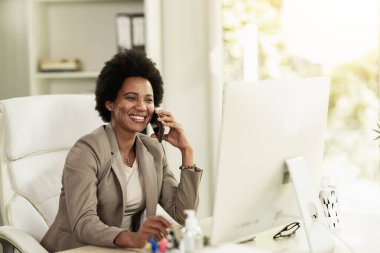 Ofiste tek başına oturup bilgisayarla uğraşırken akıllı telefondan konuşan Afrikalı Amerikalı bir iş kadını..