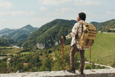 Sırt çantası ve yürüyüş direkleri olan siyah bir kadının arka görüntüsü doğada duruyor ve dağda yürüyüş yaparken temiz havanın tadını çıkarıyor..