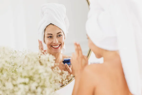 在家里的浴室里 镜子前美丽的年轻女子在脸上涂了润肤霜 — 图库照片