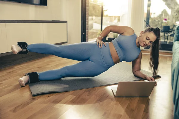 Young woman using laptop while doing plank exercises at home in the morning.