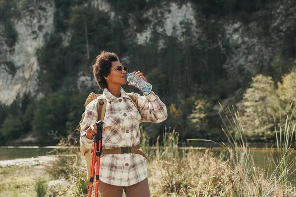 山の中でハイキング中に湖で水休憩を取るバックパック付きの熟女 — ストック写真