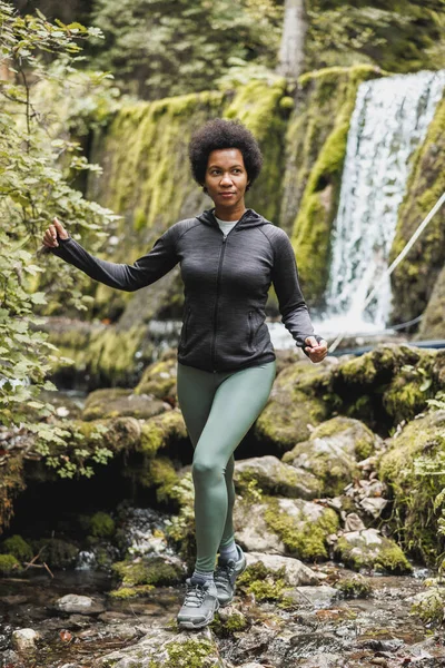 Madura Mujer Afroamericana Excursionista Cruzando Arroyo Cerca Cascada Mientras Explora —  Fotos de Stock
