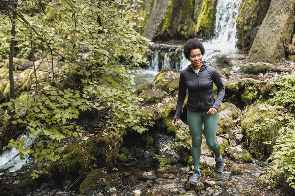 アフリカ系アメリカ人の熟女ハイカーが山の中を探検しながら滝の近くの川を渡り アウトドアトレーニングを楽しんでいます — ストック写真