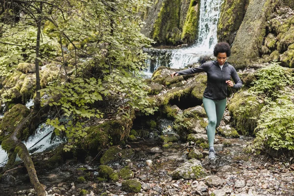 アフリカ系アメリカ人の熟女ハイカーが山の中を探検しながら滝の近くの川を渡り アウトドアトレーニングを楽しんでいます — ストック写真
