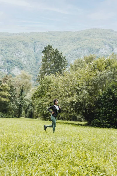 Красивая Улыбающаяся Взрослая Афроамериканка Бегает Зеленой Траве Возле Горного Ландшафта — стоковое фото