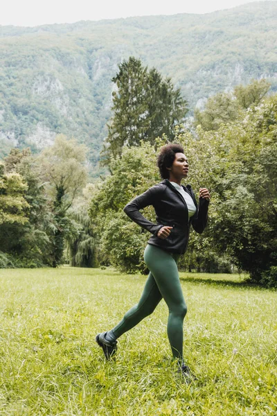 美しい笑顔成熟したアフリカ系アメリカ人女性が山の風景の近くの緑の草の中をジョギングしている — ストック写真