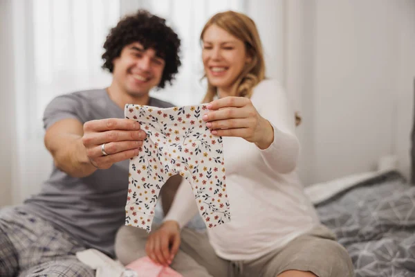 快乐的怀孕夫妇一边在卧室的床上休息一边看婴儿衣服 — 图库照片