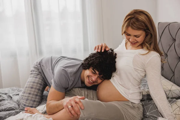 ベッドルームでリラックスしながら楽しい時間を過ごしている陽気な妊娠中のカップル 幸せな父は赤ちゃんのハートビートを聞いて — ストック写真