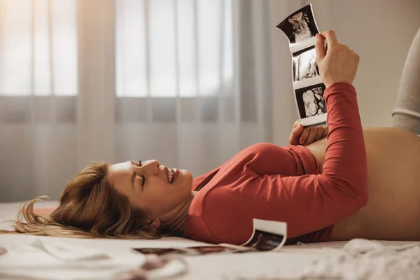 リラックスした若いです妊娠中の女性見ます超音波の彼女の赤ちゃん同時にリラックスした上でベッドルーム — ストック写真