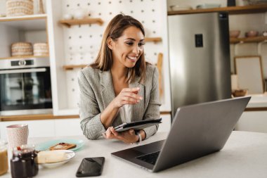 Dizüstü bilgisayar ve dijital tablet kullanan ve mutfağında kahvaltı hazırlayan çok görevli genç bir iş kadını..