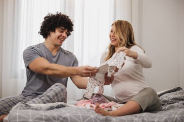 Mutlu hamile çift yatak odasında bir yatakta dinlenirken bebek kıyafetlerine bakıyor.. 