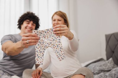 Yatak odasında bir yatakta oturan ve bebek kıyafetlerine bakan sevimli genç çift..