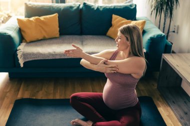 Genç hamile bir kadın sabahları evde yoga yapıyor. Oturma odasında esneme egzersizi yapıyor..