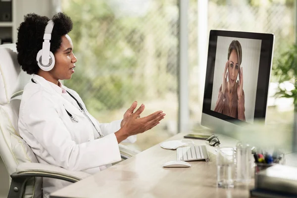 African American female doctor with headphones having video call with patient in her consulting room.