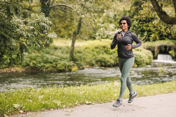 Beautiful smiling mature African American woman is jogging along trail near the mountains river in nature.