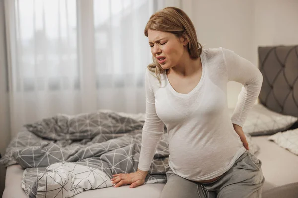 stock image Worried pregnant woman holding her back in pain while sitting on a bed in bedroom.