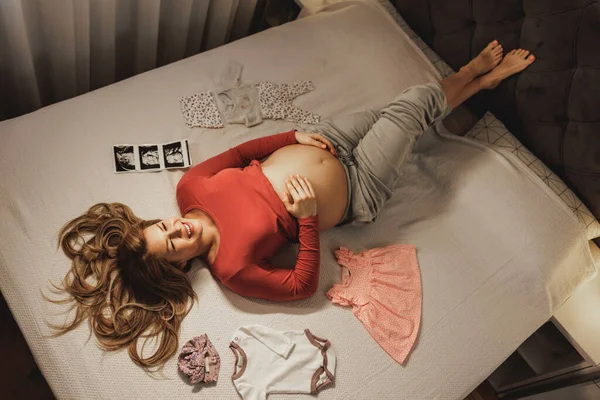 幸せな妊娠中の女性リラックスベッドの上でベッドルーム周りに赤ちゃん服 — ストック写真