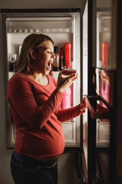夜のキッチンで開いた冷蔵庫の前でドーナツを食べる幸せな妊婦 — ストック写真
