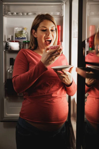 夜のキッチンのオープン冷蔵庫の前でドーナツを食べる女性を期待して幸せ — ストック写真