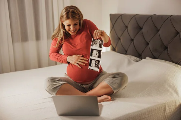 ラップトップでビデオ通話をする幸せな妊婦は ベッドルームのベッドでリラックスしながら 赤ちゃんの超音波画像を示しています — ストック写真
