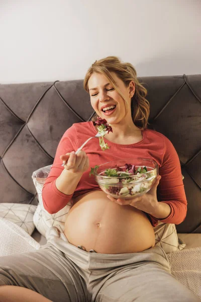 幸せな妊娠中の女性は ベッドルームのベッドに座って新鮮な野菜サラダを食べています — ストック写真